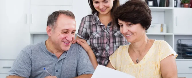 An adult daughter sits with her senior parents helping them review a memory care checklist and documents at home, representing families preparing the right questions before choosing a memory care community.