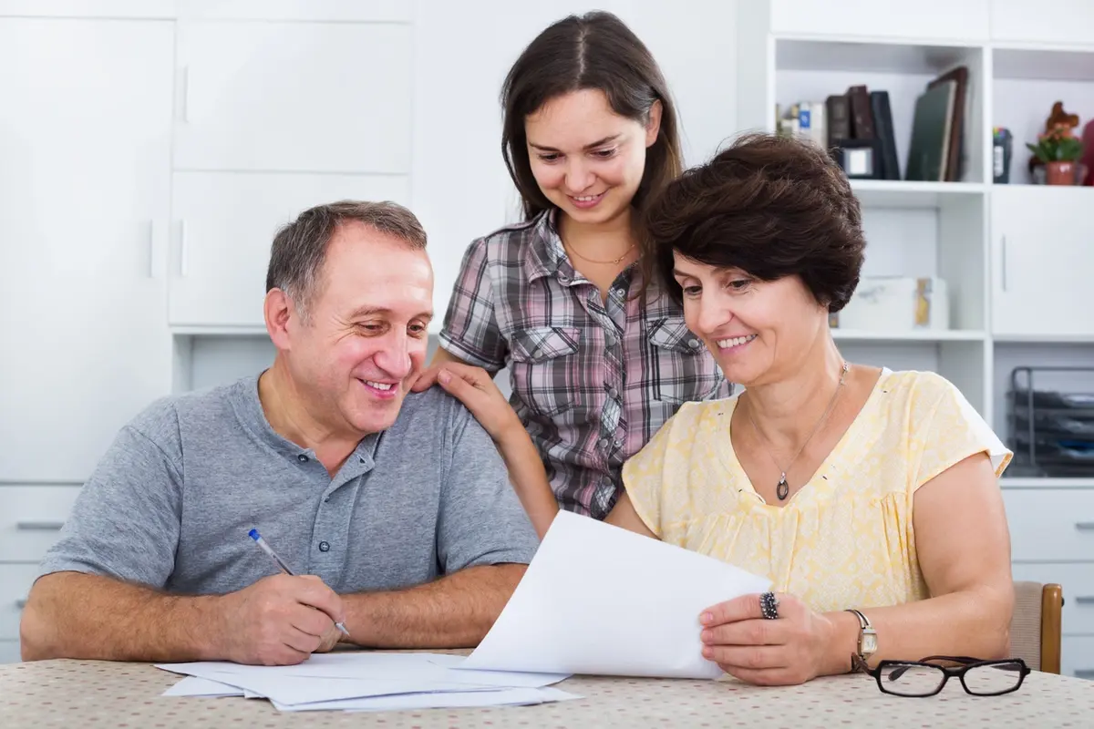 An adult daughter sits with her senior parents helping them review a memory care checklist and documents at home, representing families preparing the right questions before choosing a memory care community.